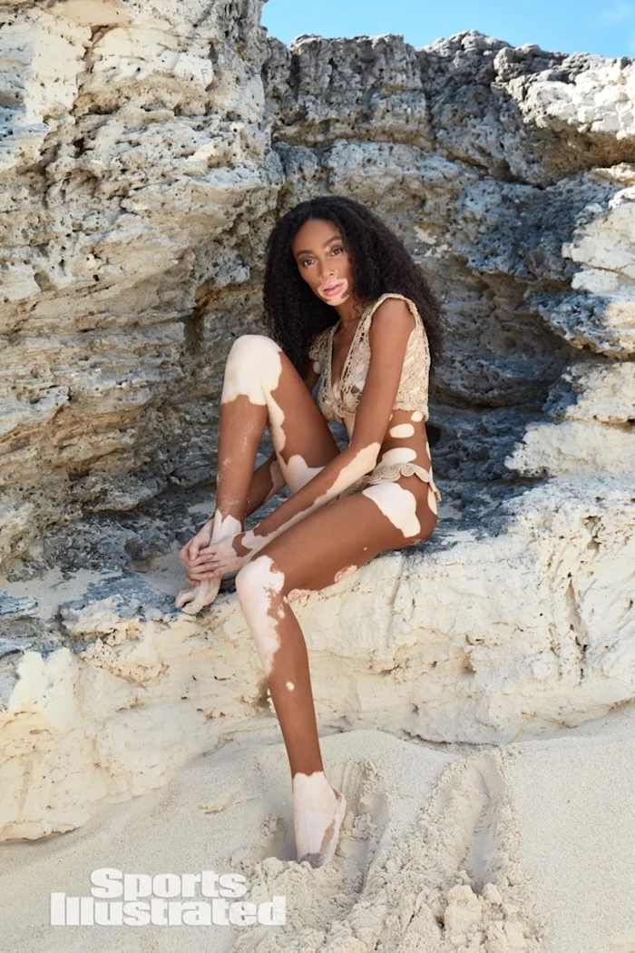 Winnie Harlow sits among the natural rock formations, wearing a beige crochet bikini.